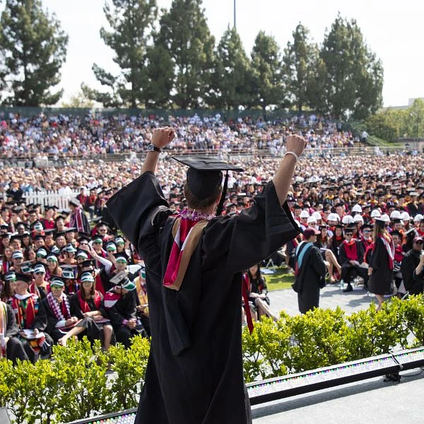 A graduate in a cap and gown raises their arms triumphantly before a large crowd at a commencement ceremony.